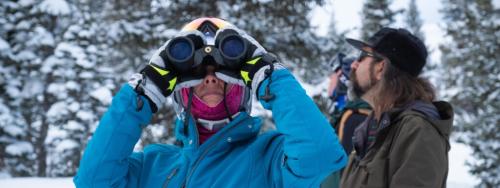 A woman in a blue coat stands in a snowy landscape and holds binoculars to her eyes.