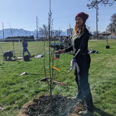 Donda holding a newly planted tree. Green grass and blue sky with mountains in the background. Donda holding a newly planted tree. Green grass and blue sky with mountains in the background.