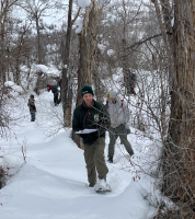 A group of people hike with snowshoes among trees in the Winter.